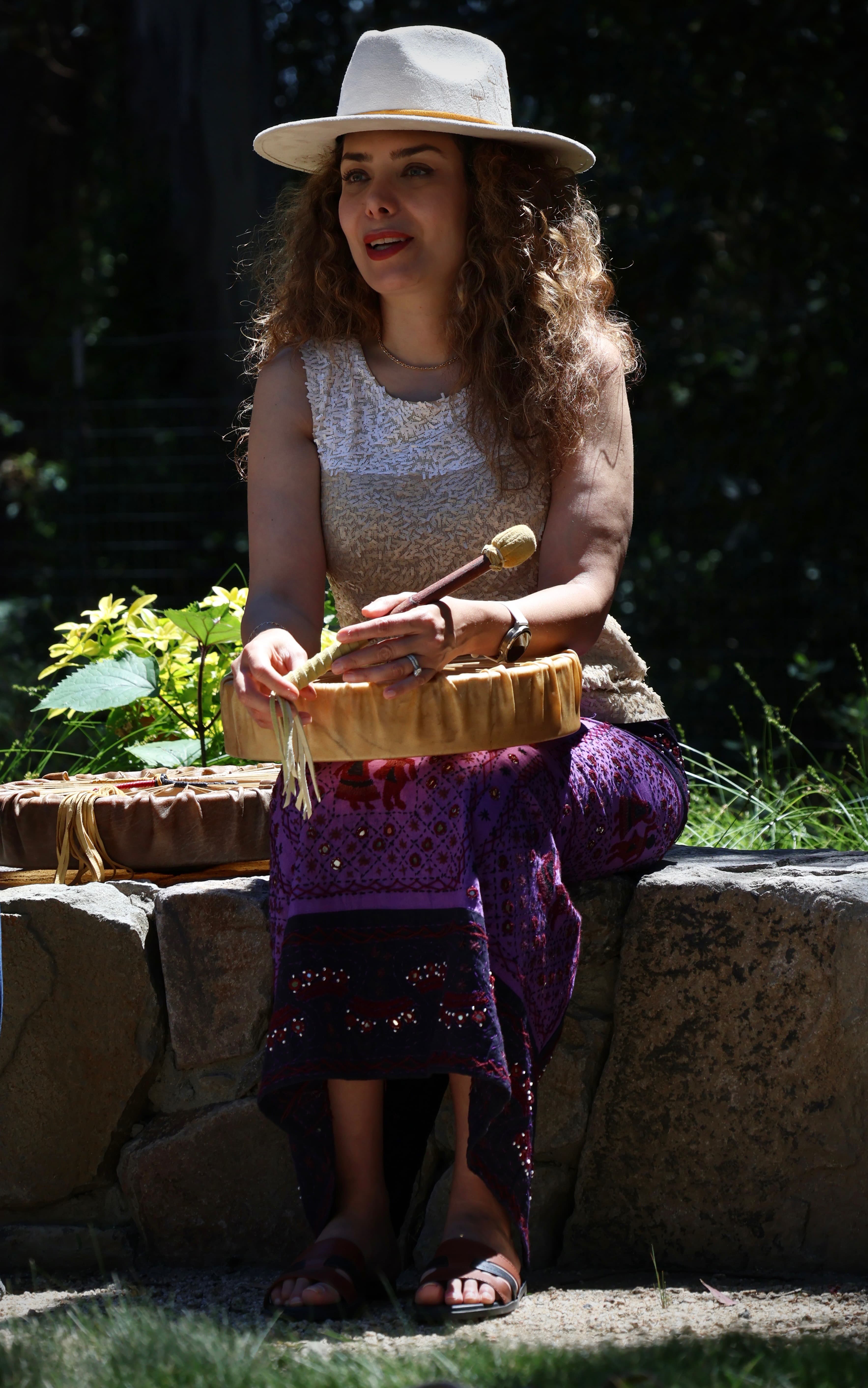 Anahita seated outdoors in a garden with a ceremonial drum